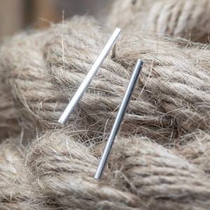 Può includere: Due orecchini a barra d'argento sono esposti su uno sfondo di corda marrone strutturata. Gli orecchini sono semplici design lineari, che riflettono la luce. Gli orecchini sono un gioiello minimalista.