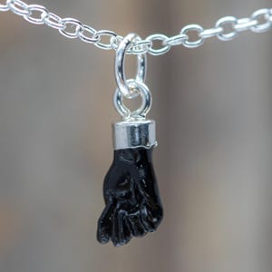 Puede incluir: Un collar de cadena plateada con un colgante en forma de mano negra en gesto de puño. El colgante está unido a la cadena mediante una argolla plateada y una tapa cilíndrica plateada en la parte superior de la muñeca, creando un contraste entre la mano negra y los componentes metálicos plateados.