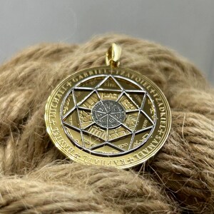 May include: A gold-colored pendant with a detailed geometric design. The pendant features a central circular element surrounded by a star-shaped frame. The outer ring has engraved text. The pendant hangs from a gold-colored bail.
