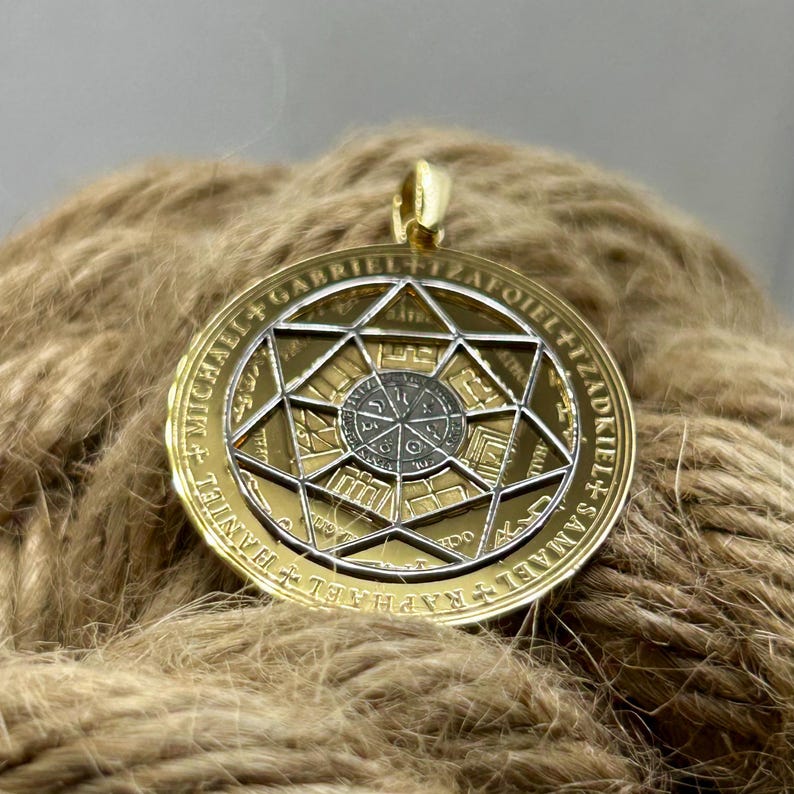 May include: A gold-colored pendant with a detailed geometric design. The pendant features a central circular element surrounded by a star-shaped frame. The outer ring has engraved text. The pendant hangs from a gold-colored bail.