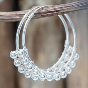 Op de afbeelding: Een paar zilveren oorringen met een cirkelvormig draadframe, versierd met een reeks kleine, gepolijste zilveren kralen die rond de onderste helft van elke ring zijn gewikkeld. De oorbellen hebben een heldere, reflecterende metalen afwerking.