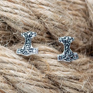 Pode incluir: Dois brincos prateados em forma de martelo de Thor. Os brincos apresentam padrões intrincados e estão colocados sobre uma superfície castanha texturizada. O design do martelo é um símbolo da mitologia nórdica.