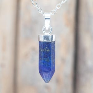 May include: A silver chain necklace with a blue lapis lazuli pendant. The pendant is a polished, elongated hexagonal shape with a silver cap. The blue stone has flecks of gold and a pointed tip.