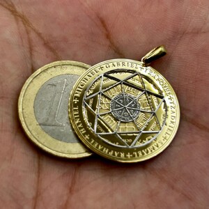 May include: A gold-colored pendant with a detailed geometric design, featuring a central star and inscribed text. The pendant is round and has a bail for a chain. A Euro coin is next to the pendant for size comparison.