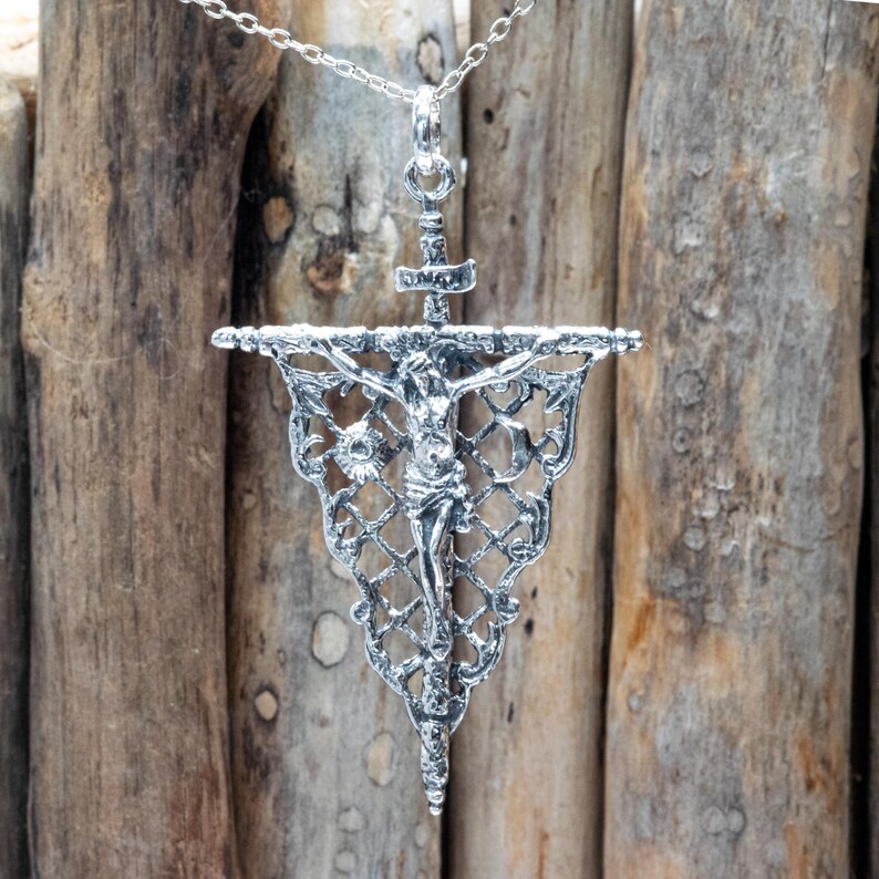 May include: Silver-toned pendant necklace featuring a detailed crucifix design. The pendant showcases a figure of Jesus on the cross, suspended from a decorative banner with intricate filigree patterns. The necklace hangs on a delicate chain.