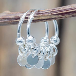 Può includere: Un paio di orecchini a cerchio in argento con ciondoli circolari pendenti. Ogni orecchino presenta una serie di piccoli dischi rotondi e piatti sospesi al cerchio. Gli orecchini sono esposti su un rametto marrone.