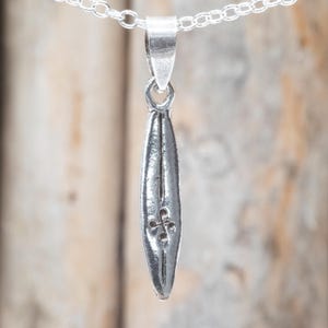 Op de afbeelding: Een zilverkleurige hangerketting met een lange, slanke, bladvormige bedel. De bedel heeft een klein, vierbladig ontwerp aan de onderkant en hangt aan een fijne zilveren ketting. De achtergrond is een vage houten ondergrond.