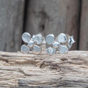 May include: A pair of silver-colored stud earrings, each shaped like a four-petal flower. The earrings are displayed on a piece of weathered wood, with a blurred wooden background.