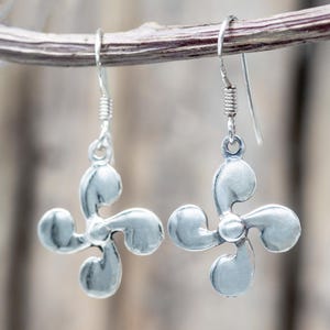 May include: A pair of silver-colored earrings. Each earring features a four-petal design with a central circular accent. The earrings hang from a simple hook and coiled wire. The earrings are displayed against a blurred, natural background.