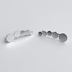 May include: A pair of silver-toned ear climbers, each featuring a series of graduated, circular discs. The discs are arranged in a curved line, creating a modern and minimalist design. The earrings are set against a plain white background.