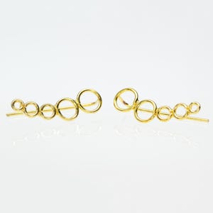 May include: A pair of gold-colored ear climbers. Each earring features a series of connected open circles, increasing in size. The earrings have a post for insertion and are displayed on a white background.