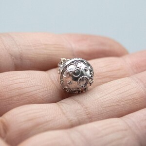 May include: A silver charm in the shape of a sphere with a textured surface and a circular design. The charm is a pendant and has a loop at the top for attaching to a chain.