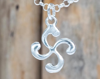 Op de afbeelding: Een zilverkleurige ketting met een hanger met een gestileerd, drielobbig ontwerp. De hanger hangt aan een fijne ketting. De achtergrond is een vage houten ondergrond.