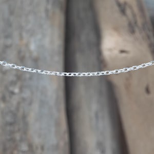 Può includere: Primo piano di una delicata catena d'argento. La catena è composta da piccole maglie ovali, creando una linea continua. Lo sfondo è sfocato, di tono neutro, evidenziando l'aspetto brillante e metallico della catena. La catena è probabilmente utilizzata per gioielli.