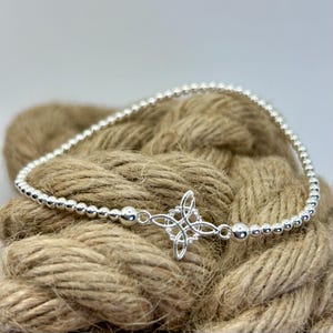 Op de afbeelding: Een zilveren kralenarmband met een centraal vierpuntig knoopornament. De armband is gemaakt van kleine, uniforme metalen bolletjes die aan elkaar zijn geregen, met een iets grotere kraal aan weerszijden van het gedetailleerde, opengewerkte knoopstuk.