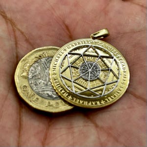 May include: A gold-colored pendant with a complex geometric design and the names of archangels inscribed around the edge. The pendant is next to a British one-pound coin. The pendant has a gold bail.