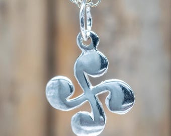 Op de afbeelding: Een zilverkleurige ketting met hanger met een Lauburu, een Baskisch symbool met vier komma-vormige elementen. De hanger hangt aan een fijne zilveren ketting. De achtergrond is wazig en neutraal.