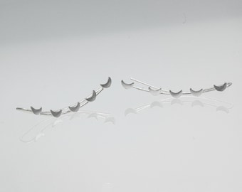 Może przedstawiać: Para srebrnych kolczyków typu ear climber, każdy z serią kształtów półksiężyca wznoszących się wzdłuż zakrzywionego pręta. Kolczyki są prezentowane na odblaskowej powierzchni, podkreślając ich delikatny design i metaliczny połysk.