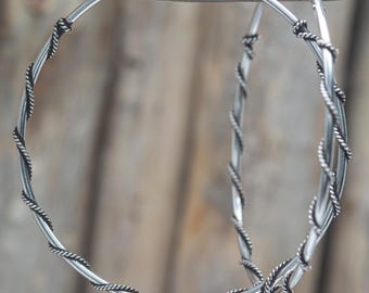 May include: A pair of large silver hoop earrings featuring a smooth circular base wrapped with a thin, twisted silver wire detail. The earrings have a polished metallic finish and a simple hinge closure mechanism at the top of each hoop.