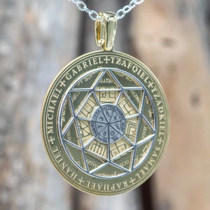 May include: A gold-toned pendant with a silver chain. The pendant features an intricate design with geometric shapes and text around the edges. The center has a detailed circular pattern. The pendant is suspended from a delicate silver chain.