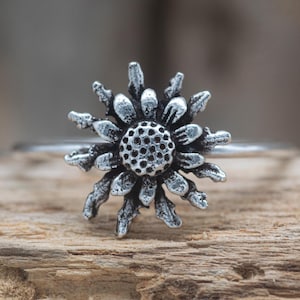 Peut inclure: Bague argentée ornée d'une fleur détaillée. La bague présente une fleur texturée avec de multiples pétales et un centre détaillé. L'arrière-plan en bois flou met en valeur le travail du métal et le thème floral.