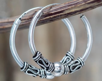 May include: A pair of silver hoop earrings with decorative detailing. The earrings are round with a small silver ball in the centre and ornate patterns on each side. The earrings are displayed on a brown twig against a blurred background.