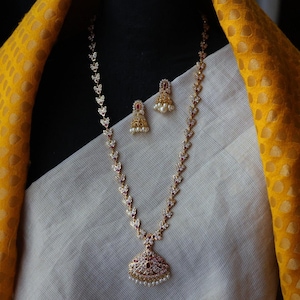 Op de afbeelding: Een goudkleurige ketting- en oorbellenset. De ketting heeft een hanger in de vorm van een druppel, versierd met kleine rode stenen en witte parels. De oorbellen hebben een vergelijkbaar ontwerp. De sieraden zijn geplaatst op een witte stof en een gele stof met patroon.