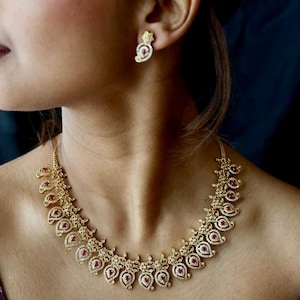 May include: A gold necklace and matching earrings set. The necklace has a repeating paisley design with red gemstones and clear crystals. The earrings have a similar design. The jewellery is worn on a person.