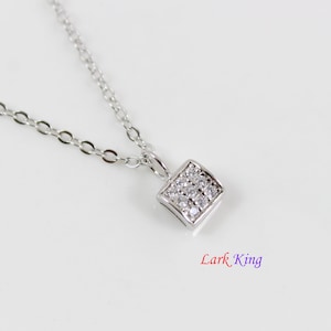 May include: A silver-toned necklace featuring a small, square-shaped pendant encrusted with multiple clear, sparkling stones. The pendant hangs from a delicate, thin silver-toned chain. The text Lark King is printed in the lower right corner.
