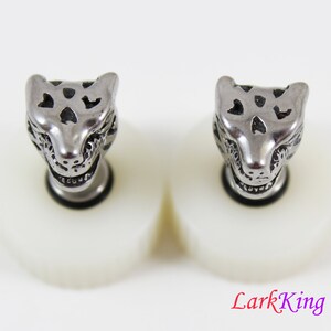 May include: A pair of silver-toned metal earrings featuring a detailed animal head design. The earrings are on a white background.