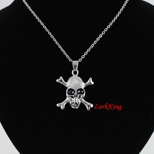 Puede incluir: Un collar con un colgante de calavera y huesos cruzados de plata. La calavera tiene ojos negros y est&aacute; detallada con una superficie texturizada. El collar est&aacute; sobre un fondo negro.