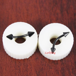May include: Two black curved barbell body piercings with pointed ends. The piercings are displayed on white circular bases.
