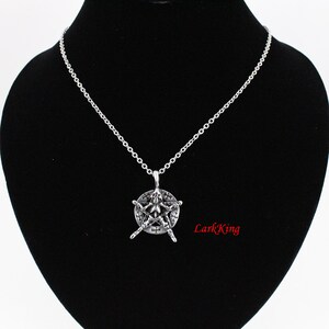 Puede incluir: Un collar de cadena de plata con un colgante que presenta una estrella de cinco puntas con un dise&ntilde;o en su interior. La estrella est&aacute; rodeada por un c&iacute;rculo con un borde elevado. El colgante est&aacute; colgado sobre un fondo de terciopelo negro.