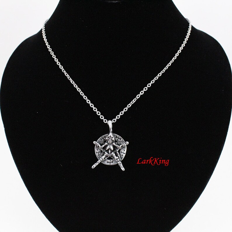 Puede incluir: Un collar de cadena de plata con un colgante que presenta una estrella de cinco puntas con un dise&ntilde;o en su interior. La estrella est&aacute; rodeada por un c&iacute;rculo con un borde elevado. El colgante est&aacute; colgado sobre un fondo de terciopelo negro.