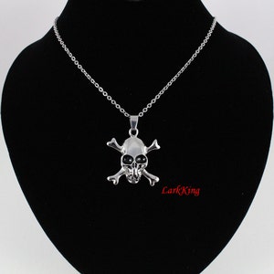 Puede incluir: Un collar de cadena de plata con un colgante de plata de calavera y huesos cruzados. La calavera tiene ojos negros.