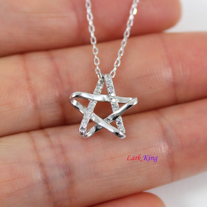 Puede incluir: Un collar con colgante de estrella plateada que presenta un dise&ntilde;o de cinco puntas, con un lado incrustado de peque&ntilde;as piedras transparentes y el otro lado con un acabado met&aacute;lico liso y pulido. El colgante cuelga de una fina cadena plateada.