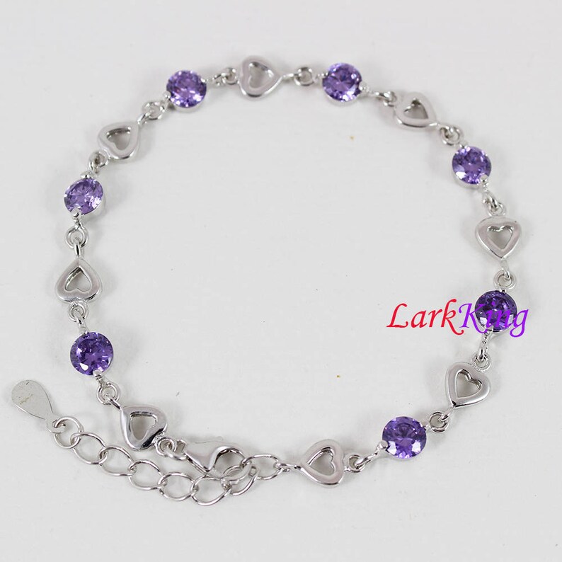 May include: A silver bracelet with a heart-shaped chain and purple gemstones. The bracelet has an adjustable clasp.