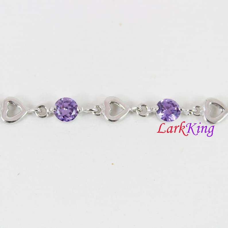 May include: A silver bracelet with a heart-shaped design and purple gemstones. Lark King is printed on the bracelet.