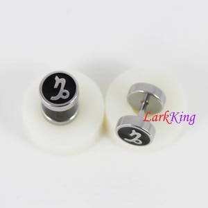 May include: A pair of silver stud earrings with black circular fronts featuring a silver Capricorn zodiac symbol.