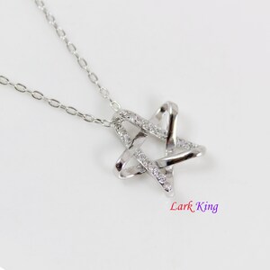 Puede incluir: Un collar de tono plateado con un colgante en forma de estrella de bandas entrelazadas. Una banda es lisa y pulida, mientras que la otra est&aacute; incrustada con peque&ntilde;as piedras claras y brillantes. El texto Lark King aparece bajo el colgante.