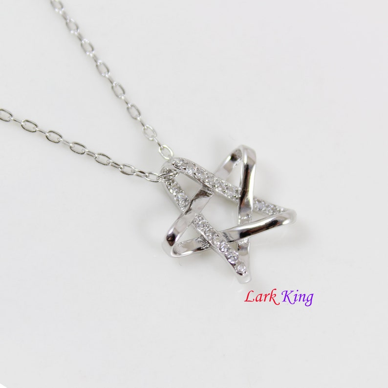 Puede incluir: Un collar de tono plateado con un colgante en forma de estrella de bandas entrelazadas. Una banda es lisa y pulida, mientras que la otra est&aacute; incrustada con peque&ntilde;as piedras claras y brillantes. El texto Lark King aparece bajo el colgante.
