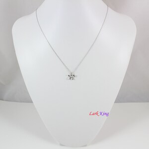 Puede incluir: Un collar de tono plateado con una cadena delicada y un peque&ntilde;o colgante de estrella de cinco puntas. El metal tiene un acabado pulido y reflectante. El texto Lark King est&aacute; impreso en la esquina inferior derecha en un degradado de rojo, azul y morado.