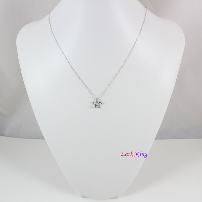 Puede incluir: Un collar de tono plateado con una cadena delicada y un peque&ntilde;o colgante de estrella de cinco puntas. El metal tiene un acabado pulido y reflectante. El texto Lark King est&aacute; impreso en la esquina inferior derecha en un degradado de rojo, azul y morado.