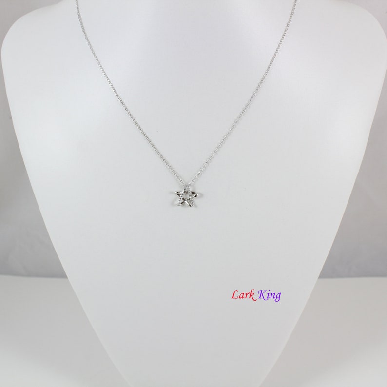 Puede incluir: Un collar de tono plateado que presenta una cadena delicada y un peque&ntilde;o colgante de estrella de cinco puntas. La estrella tiene una superficie pulida y reflectante. El texto "Lark King" est&aacute; impreso en una fuente degradada en la parte inferior derecha.