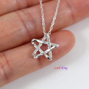 Puede incluir: Un collar con colgante de estrella plateada que presenta un dise&ntilde;o de cinco puntas con bandas entrelazadas, una de las cuales est&aacute; incrustada con peque&ntilde;as piedras claras. El colgante cuelga de una fina cadena plateada. El texto Lark King es visible.