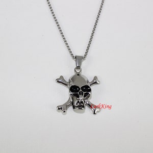 Puede incluir: Un collar de cadena de plata con un colgante de calavera y huesos cruzados. La calavera tiene ojos negros y un dise&ntilde;o detallado.