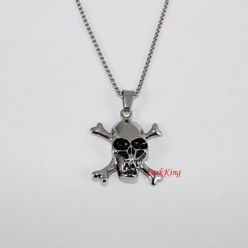 Puede incluir: Un collar de cadena de plata con un colgante de calavera y huesos cruzados. La calavera tiene ojos negros y un dise&ntilde;o detallado.