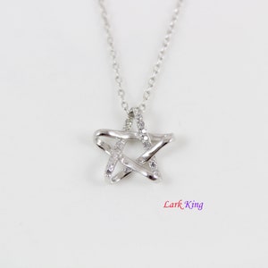 Puede incluir: Un collar de tono plateado con un colgante en forma de estrella con acabado pulido y peque&ntilde;as piedras transparentes incrustadas a lo largo de un lado de la estrella. El colgante cuelga de una fina cadena plateada. El texto Lark King es visible.