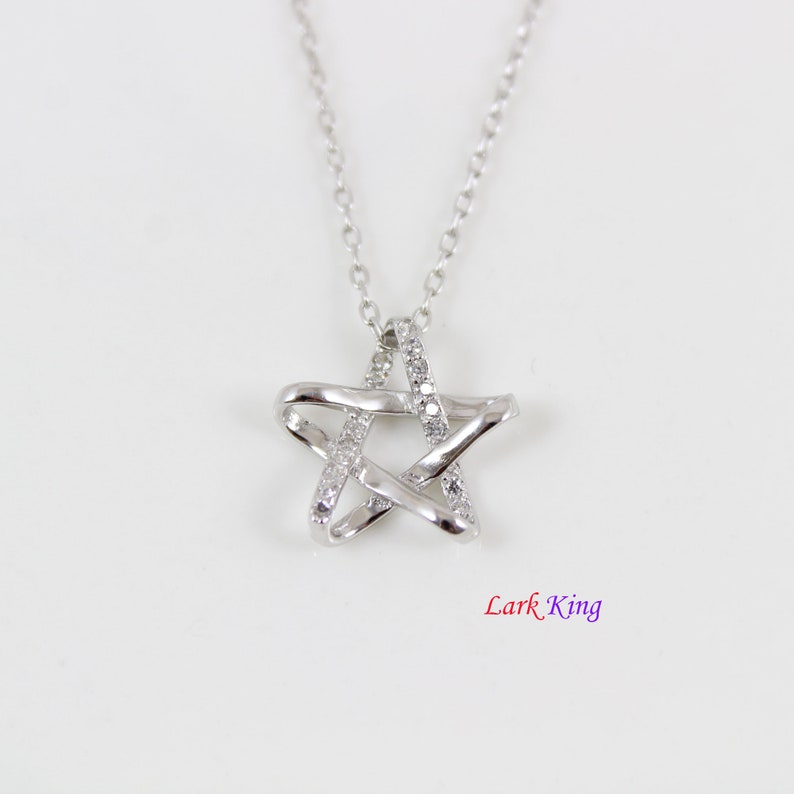 Puede incluir: Un collar de tono plateado con un colgante en forma de estrella con acabado pulido y peque&ntilde;as piedras transparentes incrustadas a lo largo de un lado de la estrella. El colgante cuelga de una fina cadena plateada. El texto Lark King es visible.