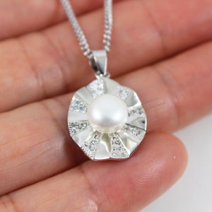Puede incluir: Un collar de plata con un colgante de gran perla blanca en el centro, rodeado de piedras pequeñas transparentes. El colgante tiene forma de flor con un diseño texturizado.